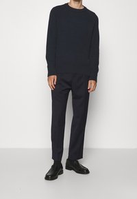 Mörk marinblå ribbad stickad tröja med rund hals, tillsammans med skräddarsydda mörka byxor och svarta slip-on skor, visad mot en enhetlig bakgrund.