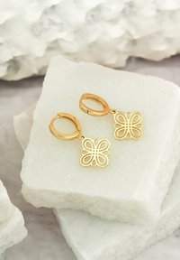 Orecchini a forma di farfalla in oro con un intricato design a intaglio, attaccati a chiusure ad anello, esposti su blocchi di pietra chiara e strutturata.