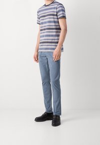 Gestreiftes Kurzarmhemd in Blau und Beige, kombiniert mit hellblauen Hosen und schwarzen Schuhen mit Kettenakzenten, an einer stehenden Figur.