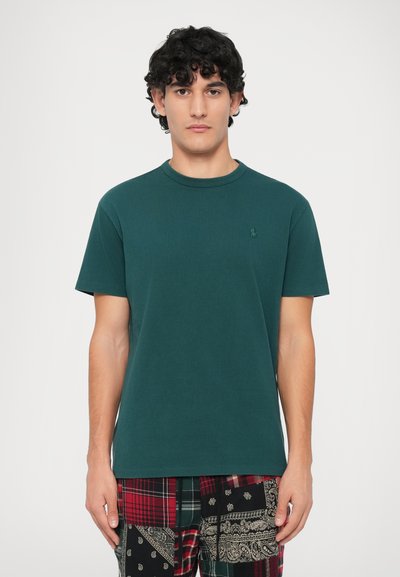 Tânăr cu păr creț și închis la culoare, îmbrăcat într-un tricou cu mânecă scurtă de culoare verde închis și pantaloni plaid din piețe colorate, stând în fața unui fundal simplu.