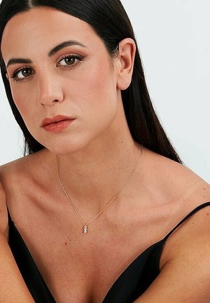 Goldene Kettenhalskette mit einem kleinen, klaren Anhänger. Das Model trägt ein schwarzes Oberteil, das das filigrane Stück vor einem neutralen Hautton zur Geltung bringt.