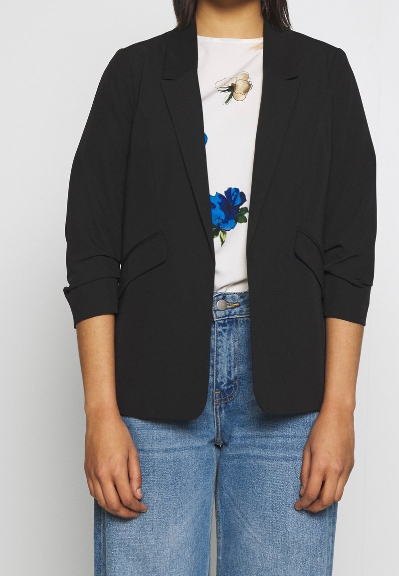 Personne portant un blazer noir sur un haut blanc à fleurs bleues et beiges, associé à un jean en denim bleu.
