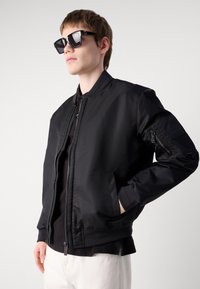 Schwarze Bomberjacke mit glatter, glänzender Oberfläche, Reißverschluss, gerippten Bündchen und Saum sowie einer Tasche am Ärmel. Getragen mit schwarzen Sonnenbrillen.