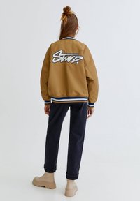 Osoba z długimi, związanymi włosami, ubrana w brązową kurtkę varsity z niebieskimi i białymi wykończeniami oraz napisem z tyłu, ciemne spodnie i beżowe buty, stojąca.