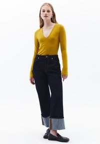 Maglietta a maniche lunghe color senape con scollo a V, abbinata a jeans in denim scuro a vita alta con risvolti azzurri chiari e righe a contrasto. Mocassini neri.
