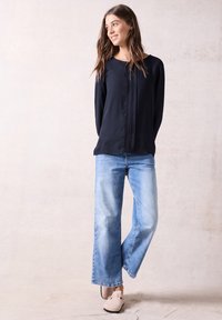 Blusa a maniche lunghe blu navy con finitura testurizzata, abbinata a jeans a gamba larga blu chiaro e scarpe slip-on beige. Design semplice e moderno.