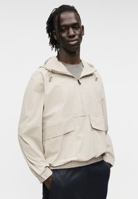 Μπεζ ελαφρύ μπουφάν με κουκούλα, φερμουάρ μπροστά και δύο μπροστινές τσέπες. Διαθέτει ελαστικά μανίκια και άνετη εφαρμογή.