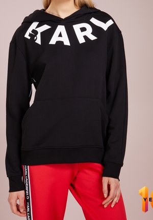 Femme portant un sweat à capuche noir avec "KARL" en lettres blanches et un pantalon rouge avec le texte "KARL LAGERFELD" sur la bande latérale.