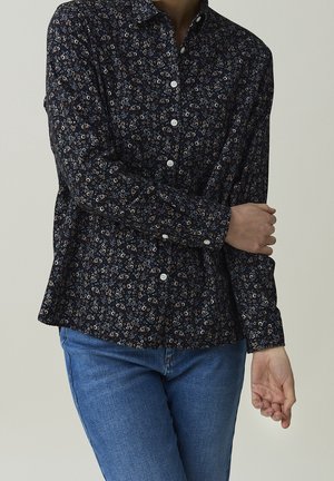 Persoon draagt een zwart shirt met lange mouwen met kleine bloemprint en witte knopen, gecombineerd met een blauwe spijkerbroek, armen gekruist ter hoogte van de taille.