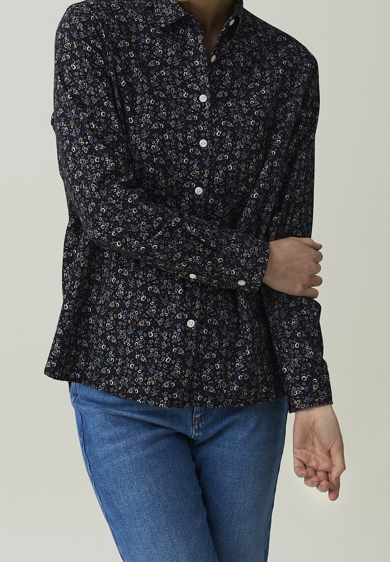 Personne portant une chemise noire à manches longues avec un petit imprimé fleuri et des boutons blancs, associée à un jean bleu, les bras croisés à la taille.