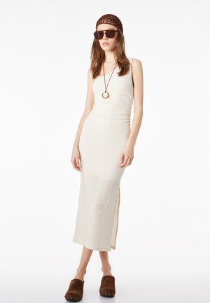 Femme en robe maxi blanche texturée sans manches, portant une casquette marron au crochet, de grandes lunettes de soleil, un long collier pendentif et des chaussures marron sans lacets.