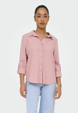 Kobieta o ciemnych włosach związanych z tyłu, ubrana w jasnoróżową koszulę zapinaną na guziki z podwiniętymi rękawami oraz jasnoniebieskie dżinsy, stojąca na tle jednolitego białego tła.