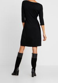 Femme portant une robe noire longue jusqu'aux genoux avec des manches trois-quarts et des bottes noires à talons hautes montantes, se tenant devant un fond uni.