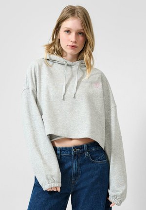 Młoda kobieta o blond włosach nosi jasnoszarą krótką bluzę z kapturem i ciemnoniebieskie jeansy z wysokim stanem, stoi na tle jednolitego szarego tła.