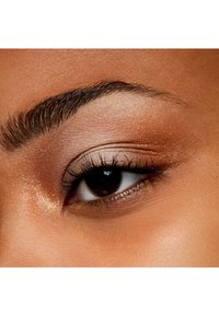 Gros plan d'un seul œil avec un iris marron foncé, des cils bien définis, un doux fard à paupières doré et un teint lisse et homogène autour.