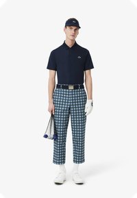 Marinblå pikétröja med Lacoste-logotyp, rutiga golfbyxor i blått och vitt, vita skor och bärande golfbollar i en nätpåse.