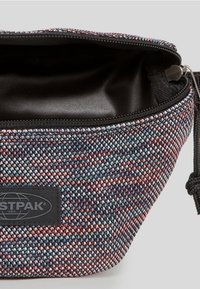 Pouzdro z tkané látky v mnohobarevném růžovém, modrém a černém vzoru. Má zapínání na zip a černou logo štítek na přední straně.