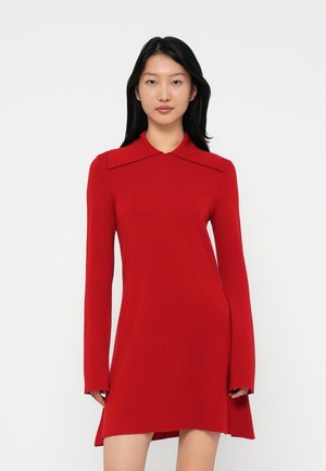 Woman with long black hair wearing a fitted red long-sleeve dress with a wide collar, standing against a white background.
