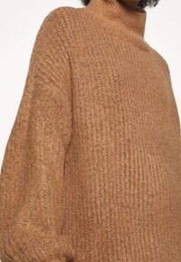 Maglione a maglia color ruggine con collo alto. Texture a coste e vestibilità oversized, con maniche ampie e una finitura morbida e pelosa.