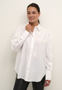 Camicia bianca con ricami a foglia, maniche lunghe e colletto classico, abbinata a pantaloni neri aderenti.