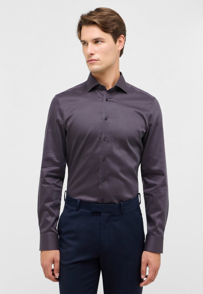 Camicia a maniche lunghe, aderente, di colore viola scuro con un fine motivo a pois. Presenta una texture liscia, apertura con bottoni sul davanti e colletto, abbinata a pantaloni scuri.