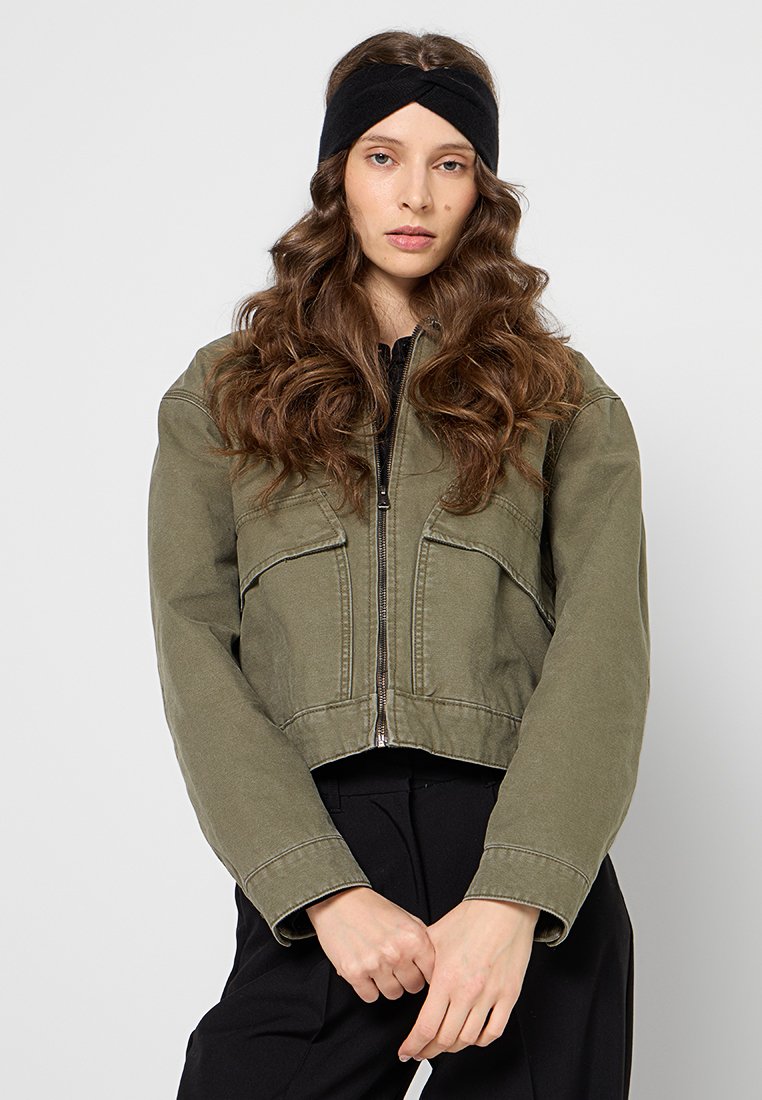 Oliwkowa kurtka z odkrytymi ramionami z zapięciem na zamek, dwoma przednimi kieszeniami i przeszyciami. Czarna opaska na głowę noszona z luźnymi, falowanymi włosami.