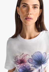 T-shirt blanc avec un motif floral dans des teintes de violet et de rose, présentant des lignes détaillées et des embellissements, col rond et manches courtes.