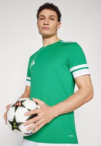 Green athletic shirt with white stripes on sleeves, made from breathable fabric. Individual holds a patterned soccer ball in front.