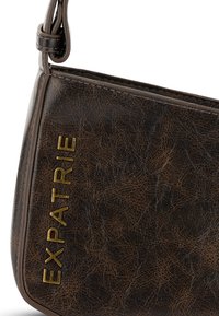 Borsa a tracolla in ecopelle marrone con testo dorato in rilievo "EXPATRIE", dalla forma strutturata e con una tracolla sottile per il trasporto a spalla.