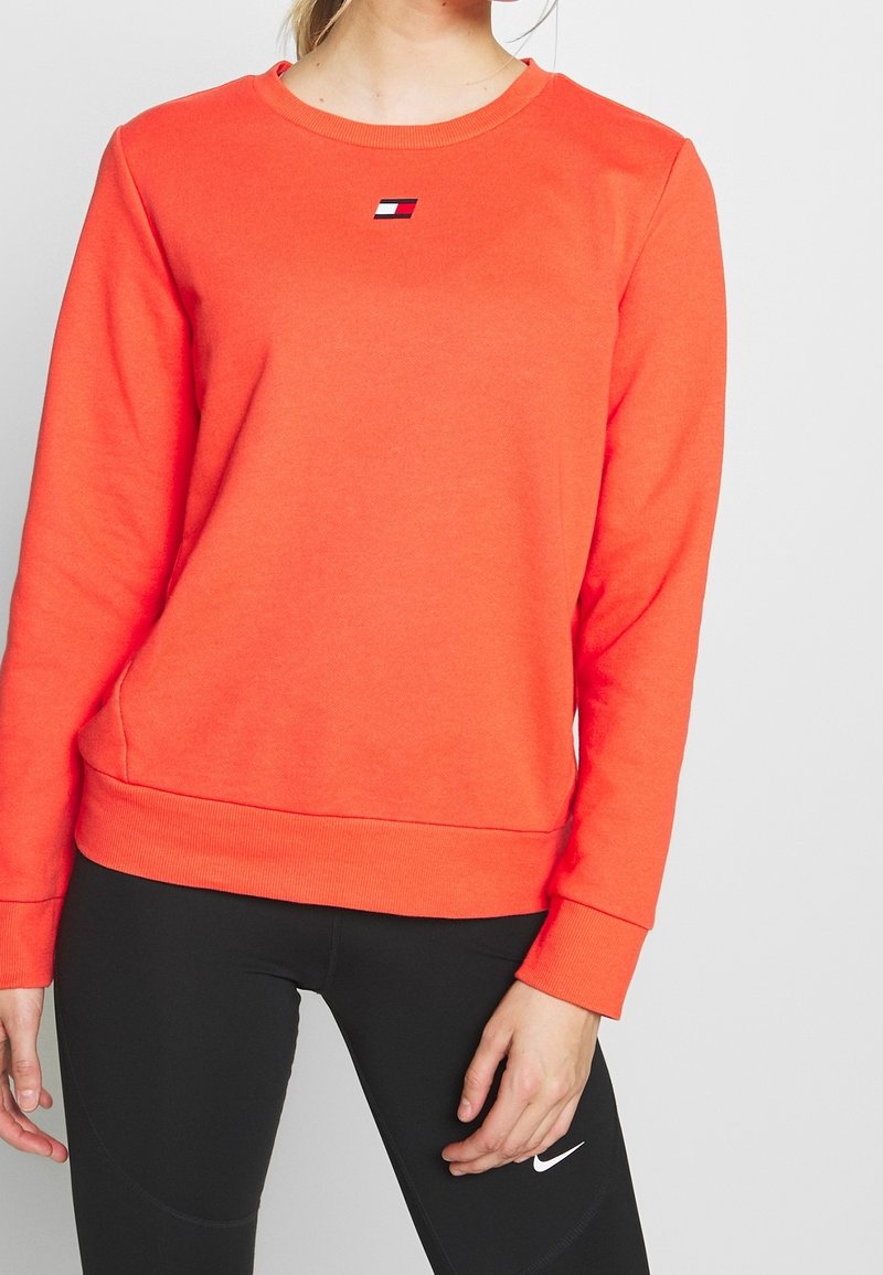 Femme portant un sweat-shirt orange vif avec un petit logo sur la poitrine et des leggings noirs avec un logo blanc sur la cuisse sur fond uni.