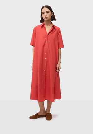 Frau mit kurzen dunklen Haaren, die ein korallenfarbenes Midi-Kleid mit Knöpfen und braune Slipper-Loafer trägt, stehend vor einem schlichten hellen Hintergrund.