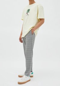 T-shirt di cotone giallo chiaro con grafica di palma e fenicottero. Pantaloni a scacchi grigi con accenti gialli e blu. Sneakers bianche con suola color caramello.