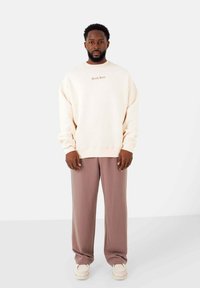 Felpa oversize color crema con un logo discreto e pantaloni beige dalla vestibilità morbida, abbinati a sneakers bianche. Texture del tessuto liscia.