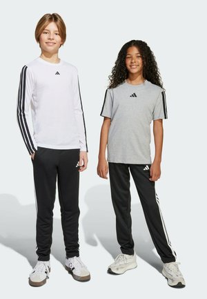 Due bambini indossano abbigliamento sportivo Adidas: una maglietta lunga bianca con strisce nere e una maglietta grigia a maniche corte, entrambe abbinate a pantaloni neri.
