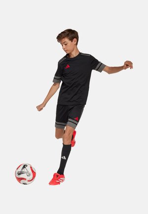 Black sports jersey with gray accents, paired with black shorts and red soccer cleats, actively kicking a red and white soccer ball.