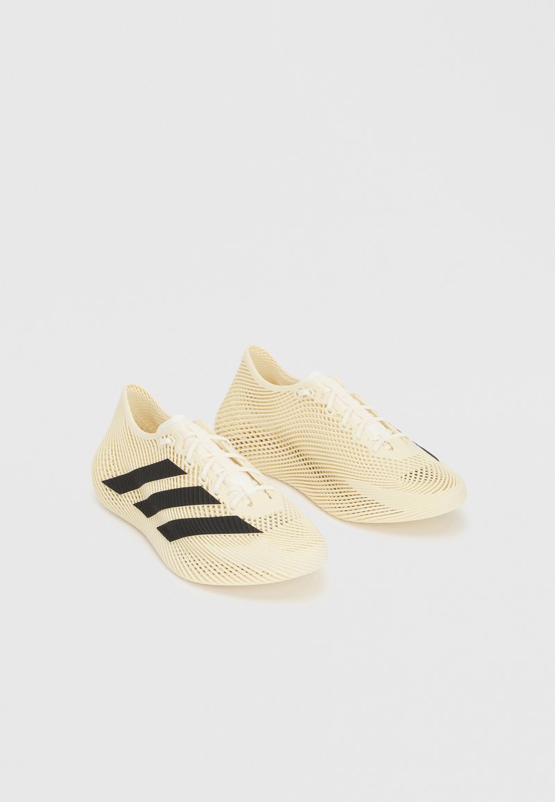 Chaussures de sport beige clair avec des bandes noires, fermeture à lacets et design texturé en forme de vagues sur fond blanc.