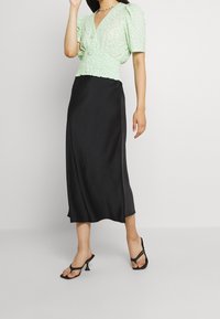 Blusa verde menta con maniche a palloncino, vita smock; gonna midi nera in satin. Tacchi neri con cinturini. Sfondo semplice.