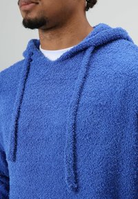 Blauwe fleece hoodie met een zachte, gestructureerde afwerking; heeft een capuchon met trekkoord en een ontspannen pasvorm met een witte binnenlaag die zichtbaar is bij de halslijn.