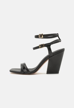 Sort højhælet sandal med en robust kilehæl, der har to tynde remme over foden og en justerbar ankelrem med gyldne spænder.