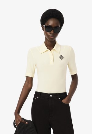 Mujer con cabello corto y rizado, lleva gafas de sol negras, polo de manga corta amarillo pálido con un pequeño logo geométrico, pantalones negros y sostiene un suéter negro.