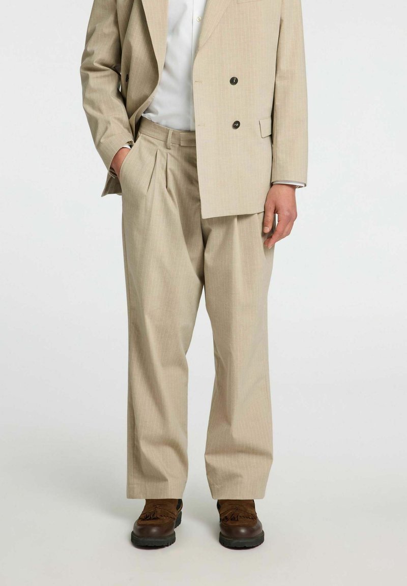 Beżowy garnitur double-breasted z subtelnym wzorem w paski, luźne spodnie oraz brązowe, fakturowane buty. Skrojony z funkcjonalnymi kieszeniami.