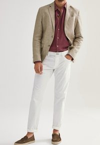 Mężczyzna w beżowej marynarce, bordowej koszuli na guziki, białych spodni z podwiniętymi nogawkami i brązowych loafersach, stoi z jedną ręką w kieszeni.