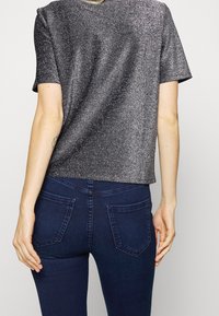 Korte mouwtop in metaalgrijze stof met een glanzende, gestructureerde afwerking, gecombineerd met donkere spijkerbroeken met twee achterzakken.