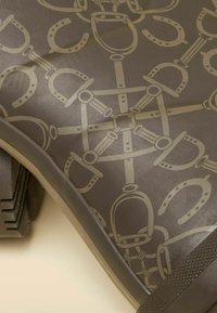 Gumené dažďové čižmy s tmavohnedým povrchom a svetlobéžovým vzorom inšpirovaným jazdeckou tematikou, vrátane strmeňov a spojov.