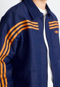 Mörkblå jacka med strukturerat mönster, orangea tre ränder på axlarna och en liten logotyp. Har en krage och dragkedjestängning.