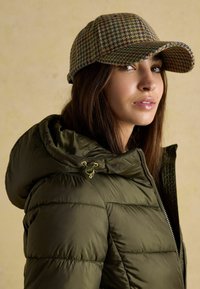 Vycpaná olivovo zelená bunda s vysokým golierom, skombinovaná s viacfarebnou kockovanou čiapkou s zakriveným okrajom a textúrovanou látkou.