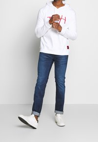 Sudadera blanca con texto gráfico rojo, jeans de mezclilla y zapatillas blancas. La sudadera cuenta con un cordón, puños y una etiqueta de logo en el lateral.