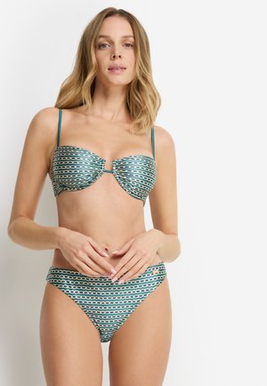 Žena s blond vlasmi po plecia, ktorá má na sebe bikiny s modrozeleným a béžovým vzorom, stojí so zľahka položenými rukami na páse.