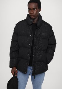 Black puffer jacket with a textured dot pattern, front buttons, and a high collar. Worn with a black shirt and blue jeans.