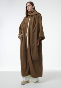 Abrigo tamaño grande, marrón y esponjoso, con un cuello alto y mangas largas, que cuenta con dos bolsillos laterales y una textura suave, colocado sobre un atuendo blanco.